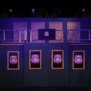 A picture of a blue and pink stage set with two levels, the rear level behind a handrail and decking section. There are four doors on the top section and two on the bottom. The set is dressed with TVs in picture frames. One large upper TV in 'landscape' orientation and four below in portrait / vertical orientation between the two lower doors